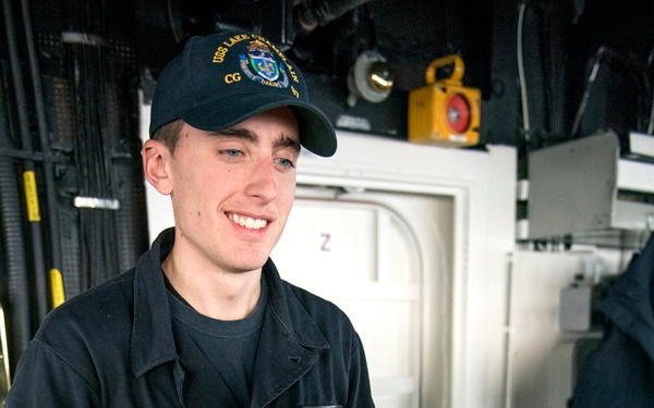A young U.S. Navy sailor in dark blue coveralls is stationed at what appears to be a ship's helm or control console aboard a naval vessel. He is wearing a cap bearing the text 'USS Lake Champlain CG' and is smiling while operating controls. The setting is the bridge or interior control room of a U.S. Navy guided-missile cruiser.