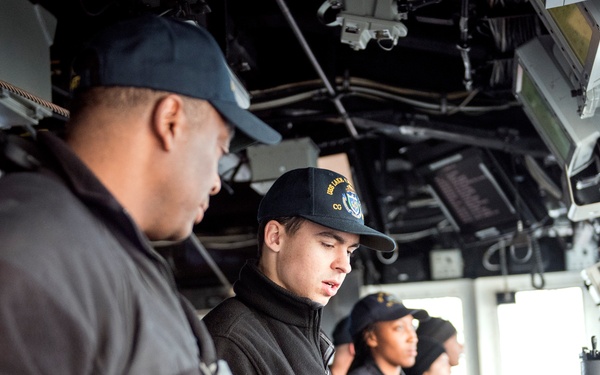 The image shows several naval personnel on the bridge of a military vessel, engaged in navigation and watch duties. Personnel are wearing dark navy uniforms and caps, with at least one cap bearing ship insignia. The bridge is equipped with navigation consoles, monitors, binoculars, and overhead equipment typical of a U.S. Navy warship.