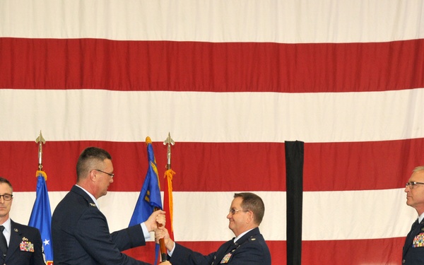 A military change of command ceremony is taking place inside what appears to be an aircraft hangar. Two Air Force officers are exchanging a guidon (unit flag), which is the central symbolic act of a change of command ceremony. Several other uniformed personnel are present on stage, with a large American flag serving as the backdrop.