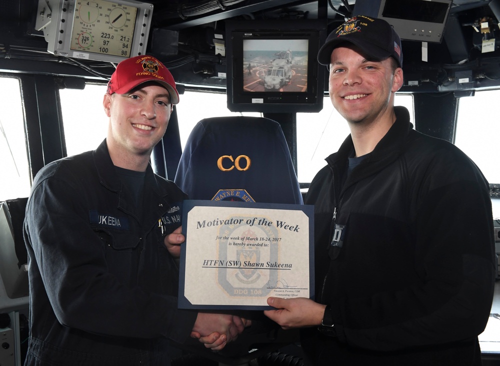 Two U.S. Navy personnel are posing on the bridge of a naval vessel, holding a 'Motivator of the Week' certificate awarded to HTFN (SW) Shawn Sukeena for the week of March 18-24, 2017. The ceremony appears to take place aboard DDG-108, with the Commanding Officer's chair visible in the background. Both individuals are smiling and shaking hands while presenting the award.