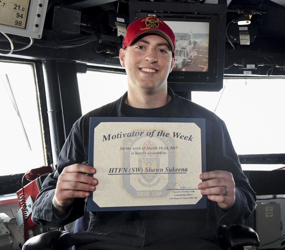 A smiling young sailor aboard USS Wayne E. Meyer (DDG 108) holds a 'Motivator of the Week' certificate awarded for the week of March 18-24, 2017. The certificate is awarded to HTFN (SW) Shawn Sukeena and is signed by Commanding Officer Vincent A. Forston, CDR. The photo appears to be taken on the ship's bridge, surrounded by navigation equipment and monitors.