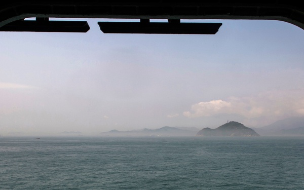 A view through a large rectangular porthole or ship window looking out over open sea toward a cluster of islands in the distance. The water is a blue-green color with light chop, and the atmosphere is hazy with partial cloud cover. A small vessel is faintly visible on the far left horizon.