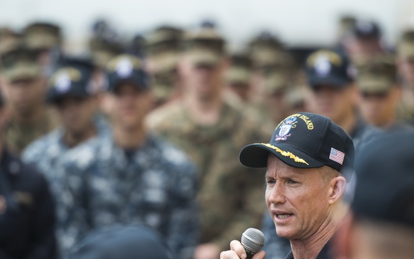 A senior naval officer is addressing a large group of military personnel while holding a microphone. He is wearing a dark navy uniform with a name tag and a black baseball cap bearing an American flag patch and what appears to be a ship's insignia. The audience consists of sailors and soldiers in various uniforms, gathered in formation to listen to the speaker.