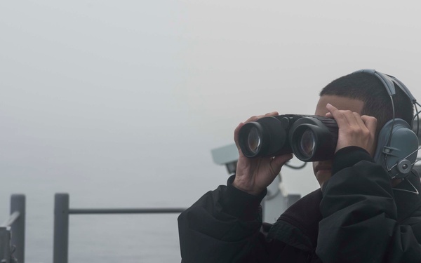 A person wearing a black jacket and communication headset stands on the deck of a naval vessel, scanning the horizon through binoculars in foggy conditions. A red 'DANGER AREA' warning sign is visible in the foreground, along with ship railings and equipment. The overcast, misty weather significantly reduces visibility over the open water.