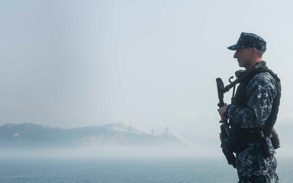 A uniformed military personnel, appearing to be a U.S. Navy sailor in digital blue camouflage (NWU Type I), stands at attention on the deck of a naval vessel while holding a rifle. The sailor is equipped with tactical gear including a vest and backpack, standing watch over a body of water with a fog-shrouded island visible in the background. The scene suggests a security watch or port approach detail.