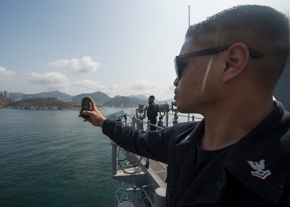 A U.S. Navy sailor, identifiable by the eagle rating badge on his uniform, stands on the deck of a naval vessel holding a handheld device — likely a GPS or radio — extended outward. A second crew member is visible in the background operating equipment on the ship's bridge wing. The vessel appears to be navigating coastal waters with mountainous terrain and urban structures visible in the distance.