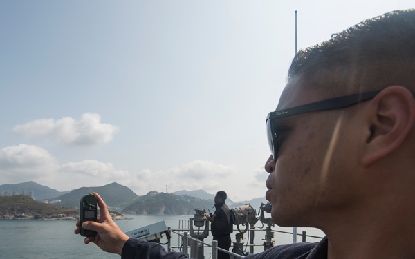A U.S. Navy sailor, identifiable by the eagle rating badge on his uniform, stands on the deck of a naval vessel holding a handheld device — likely a GPS or radio — extended outward. A second crew member is visible in the background operating equipment on the ship's bridge wing. The vessel appears to be navigating coastal waters with mountainous terrain and urban structures visible in the distance.