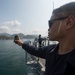 A U.S. Navy sailor, identifiable by the eagle rating badge on his uniform, stands on the deck of a naval vessel holding a handheld device — likely a GPS or radio — extended outward. A second crew member is visible in the background operating equipment on the ship's bridge wing. The vessel appears to be navigating coastal waters with mountainous terrain and urban structures visible in the distance.