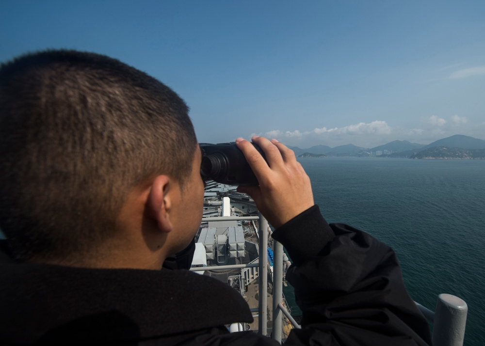 A person in a black jacket is using binoculars while standing on an elevated position aboard what appears to be a naval vessel. The individual is looking out toward a body of water with mountainous coastline and a distant urban area visible on the horizon. The ship's deck equipment and superstructure are visible below the observer's vantage point.