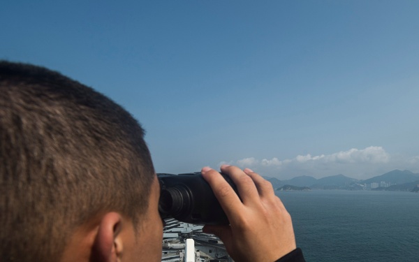 A person in a black jacket is using binoculars while standing on an elevated position aboard what appears to be a naval vessel. The individual is looking out toward a body of water with mountainous coastline and a distant urban area visible on the horizon. The ship's deck equipment and superstructure are visible below the observer's vantage point.