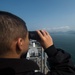 A person in a black jacket is using binoculars while standing on an elevated position aboard what appears to be a naval vessel. The individual is looking out toward a body of water with mountainous coastline and a distant urban area visible on the horizon. The ship's deck equipment and superstructure are visible below the observer's vantage point.