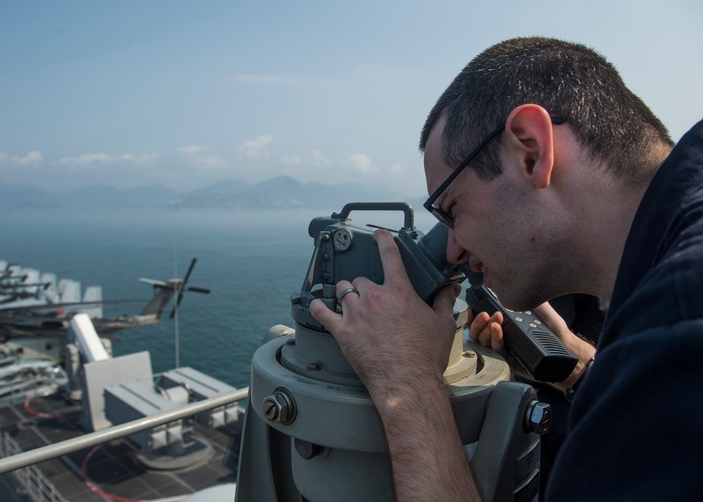 A male sailor wearing glasses and a dark uniform is looking through a bearing circle or alidade mounted on a ship's bridge wing. The vessel appears to be a naval warship, likely an aircraft carrier or large surface combatant, positioned near a coastline with mountains visible in the background. The sailor is also holding a radio handset, suggesting he is conducting navigation or watchstanding duties.