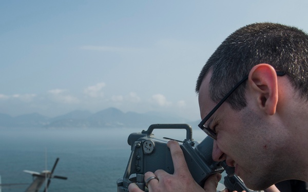 A male sailor wearing glasses and a dark uniform is looking through a bearing circle or alidade mounted on a ship's bridge wing. The vessel appears to be a naval warship, likely an aircraft carrier or large surface combatant, positioned near a coastline with mountains visible in the background. The sailor is also holding a radio handset, suggesting he is conducting navigation or watchstanding duties.