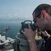 A male sailor wearing glasses and a dark uniform is looking through a bearing circle or alidade mounted on a ship's bridge wing. The vessel appears to be a naval warship, likely an aircraft carrier or large surface combatant, positioned near a coastline with mountains visible in the background. The sailor is also holding a radio handset, suggesting he is conducting navigation or watchstanding duties.