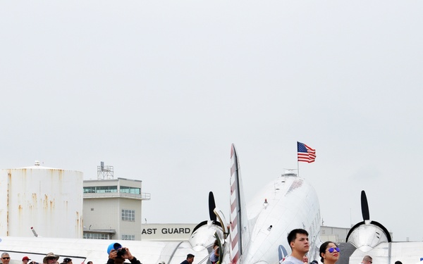 A large crowd of spectators is gathered at what appears to be a Coast Guard air show or aviation event. Attendees are seated in folding chairs and standing on a tarmac, watching aerial performances while a vintage twin-engine aircraft is displayed in the background. An American flag is visible flying near the aircraft, and Coast Guard facility buildings are seen in the background.