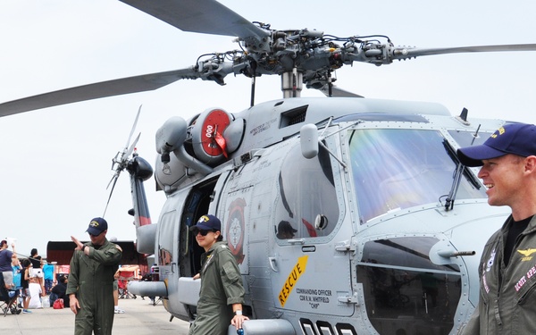 A military helicopter, identified as a rescue aircraft with tail number '000', is on display at what appears to be an air show or public military event. Several personnel in olive green flight suits and navy blue caps are standing near the helicopter, interacting with visitors. The commanding officer's name 'CDR R.H. WHITFIELD' and the word 'RESCUE' are visible on the fuselage.