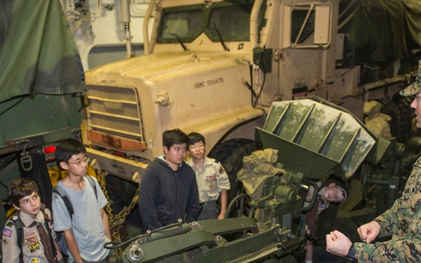 A U.S. Marine in digital camouflage uniform is briefing a group of Boy Scouts and youth visitors about military equipment inside what appears to be a military vessel or hangar. The group is gathered around heavy military hardware including what appears to be an artillery piece or weapons system, with a USMC-marked military truck visible in the background. The setting suggests an educational tour or outreach event.