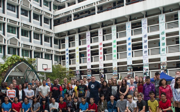 A large group of approximately 60-80 people, appearing to be a mix of Asian and Western individuals of various ages, pose for a group photo on an outdoor basketball court. The setting appears to be a school or educational institution in a Chinese-speaking region, likely Hong Kong, based on the traditional Chinese characters displayed on vertical banners. The gathering suggests a cultural exchange, school visit, or international program event.
