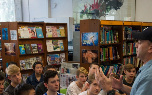 A man wearing a black cap and blue shirt is speaking and gesturing to a group of young people seated in what appears to be a school or public library. The audience consists of approximately 15-20 teenagers and at least one adult, all attentively listening to the presenter. The setting suggests an educational talk, author visit, or informational session.