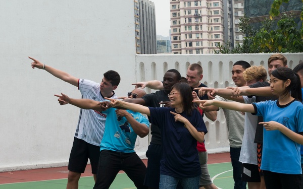 A diverse group of approximately 10 people are gathered on an outdoor basketball court, all pointing in the same direction in a coordinated pose. The group appears to be participating in a team-building or group activity, likely in an urban Asian setting based on the surrounding architecture. The atmosphere is playful and energetic, with participants of various ages and ethnicities engaged together.