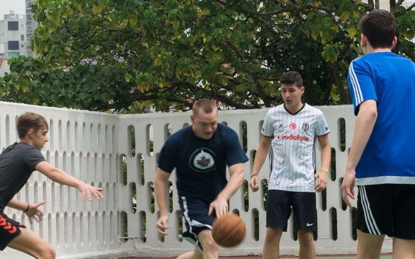 Four young men are playing basketball on an outdoor court with a green surface. One player is actively dribbling the ball while others are in defensive or observational stances. The setting appears to be an urban recreational court surrounded by a decorative white wall and trees.