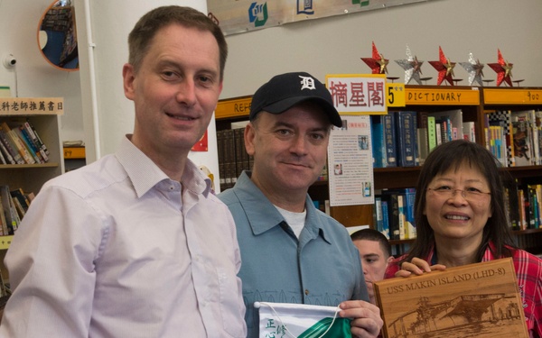 Three individuals are posing together in what appears to be a school library with Chinese-language signage, likely in Hong Kong. They are holding a commemorative plaque engraved with 'USS Makin Island (LHD-8)', a USS Makin Island baseball cap, and a school pennant/flag. This appears to be a ceremonial exchange or goodwill visit between U.S. Navy personnel and a local school.