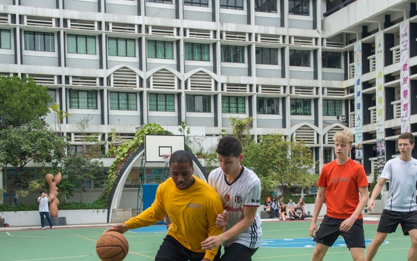 A basketball game is being played on an outdoor court surrounded by a multi-story building with Chinese-language banners visible on the right side, suggesting an Asian urban setting, likely Hong Kong or mainland China. Several young men of mixed ethnicities are actively playing, with one player in a yellow jersey dribbling while being defended. Spectators are seated in the background near a blue canopy tent.