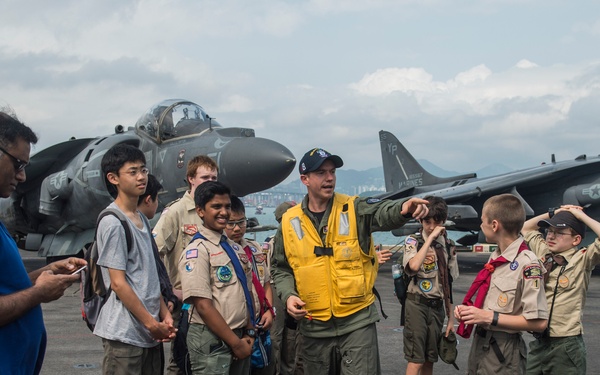 A military personnel member wearing a yellow safety vest is giving a guided tour to a group of Boy Scouts on what appears to be the flight deck of a US Navy/Marine Corps vessel. Several AV-8B Harrier jump jets with 'MARINES' markings are visible in the background. The group includes youth in Boy Scout uniforms accompanied by adult chaperones.