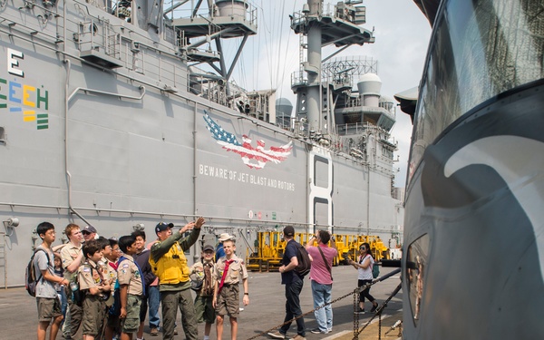A group of Boy Scouts and adult chaperones are receiving a guided tour on the flight deck of a U.S. Navy amphibious assault ship. A uniformed Navy or Marine Corps personnel member is gesturing and explaining something to the group. A military helicopter (partially visible in the foreground) is chained to the deck, and the ship's island superstructure displays an American eagle graphic with the warning 'BEWARE OF JET BLAST AND ROTORS.'