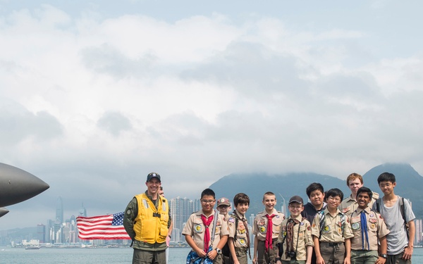 A group of Boy Scouts and civilians pose for a photo on the flight deck of a US Navy aircraft carrier with a naval aviator wearing a yellow flight vest. An American flag is visible being held in the background, and a city skyline with mountains — consistent with Hong Kong's Victoria Harbour — is visible in the distance. A military aircraft with the number '55' is partially visible on the left side of the frame.