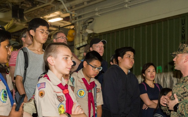 A U.S. Marines service member in digital camouflage uniform is briefing a group of Boy Scouts and their accompanying adults inside what appears to be the interior of a military vessel or facility. Several scouts are wearing the traditional tan Boy Scouts of America uniforms with neckerchiefs, patches, and merit badges visible. The setting appears to be an official tour or educational visit to a military installation.