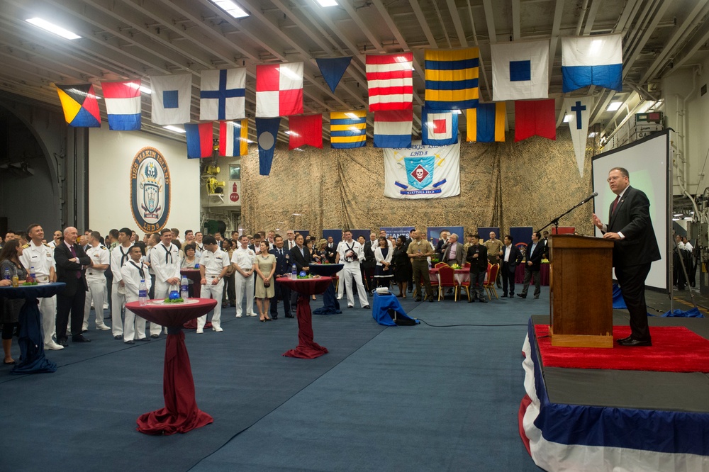 A formal military ceremony or reception is taking place aboard what appears to be a U.S. Navy ship, likely the USS Makin Island (LHD-8) based on the visible ship insignia on the left wall. A man in a dark suit is speaking at a wooden podium on a raised red-carpeted stage, addressing a large audience of military personnel in dress uniforms and civilians. The hangar bay is decorated with international maritime signal flags and unit insignia banners.