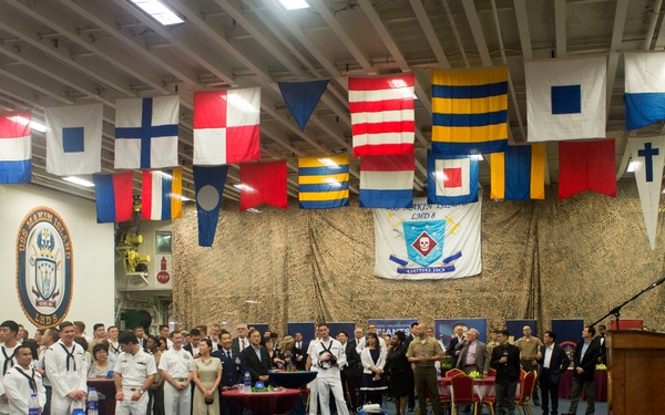 A formal military ceremony or reception is taking place aboard what appears to be a U.S. Navy ship, likely the USS Makin Island (LHD-8) based on the visible ship insignia on the left wall. A man in a dark suit is speaking at a wooden podium on a raised red-carpeted stage, addressing a large audience of military personnel in dress uniforms and civilians. The hangar bay is decorated with international maritime signal flags and unit insignia banners.