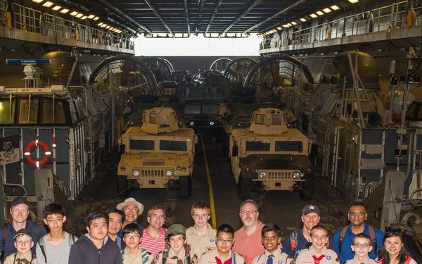 A group of Boy Scouts and adult chaperones pose for a group photo inside the vehicle deck of a large military naval vessel, likely a amphibious transport or landing ship. Several military Humvees are parked in rows behind the group, illuminated by overhead lighting. The setting appears to be an organized tour or educational visit to the ship.