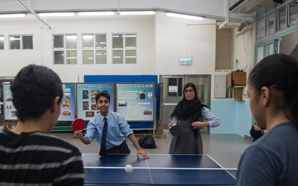 A group of people are playing table tennis in what appears to be a school gymnasium or recreational hall. Two players on one side face two players on the opposite side, with a ping pong ball visibly in motion above the net. The setting includes bulletin boards with Chinese text and posters in the background.