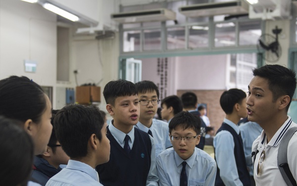 A group of students in school uniforms are gathered in what appears to be a school hall or common room, engaged in conversation with a young man wearing a white polo shirt and backpack. The uniformed students are wearing light blue shirts with dark ties and vests, suggesting a formal school dress code. The setting appears to be an educational institution, possibly during an event, orientation, or informal discussion session.