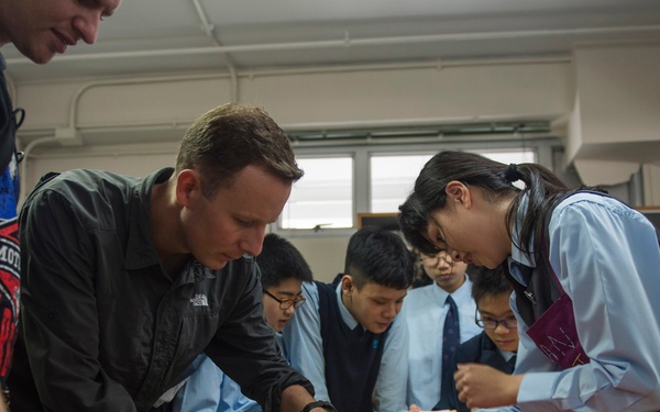 A group of students in school uniforms (light blue shirts and ties) are gathered around a wooden table in what appears to be a home economics or cooking classroom. Two Western adults are participating alongside the students in a hands-on food preparation activity. A female student wearing a purple apron appears to be demonstrating or leading the activity.