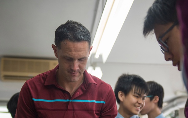 A man in a red striped polo shirt is handling a small piece of paper or food wrapper at a table with food items, surrounded by students in school uniforms. The setting appears to be a classroom or school cafeteria environment. The students, wearing dark blazers with ties and light blue shirts, are observing or participating in what appears to be a food-related activity or demonstration.