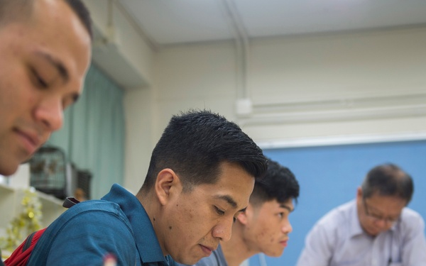 A classroom scene showing multiple male students seated at desks, actively writing or drawing on paper. An older male figure, likely an instructor or teacher, is visible in the background leaning over a desk. The image is taken from a low angle, giving a close-up perspective of the students at work.