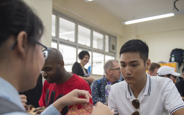 A diverse group of people are gathered in what appears to be a classroom setting, engaged in a hands-on craft or paper-folding activity. A young Asian male in a white polo shirt watches attentively as a person in a light blue shirt works with colorful patterned paper. The scene suggests a cultural exchange or workshop event with participants of various backgrounds.