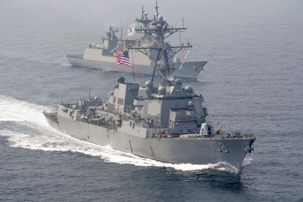 The image shows a formation of naval warships underway at sea, with a U.S. Navy Arleigh Burke-class guided-missile destroyer bearing hull number 108 in the foreground. Behind it are at least two additional warships, one of which displays a South Korean flag, suggesting a joint naval exercise or patrol operation. The ships are moving at speed, generating visible bow waves and wakes.