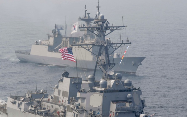 The image shows a formation of naval warships underway at sea, with a U.S. Navy Arleigh Burke-class guided-missile destroyer bearing hull number 108 in the foreground. Behind it are at least two additional warships, one of which displays a South Korean flag, suggesting a joint naval exercise or patrol operation. The ships are moving at speed, generating visible bow waves and wakes.