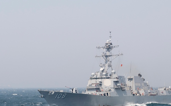 A U.S. Navy Arleigh Burke-class guided-missile destroyer bearing hull number 108 is underway at sea in moderately rough conditions. The vessel is traveling at speed, generating a visible bow wake and stern wake. The sky is overcast and hazy, suggesting reduced visibility conditions.