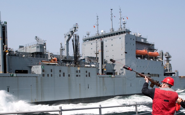 Two naval personnel wearing red helmets and red vests are conducting an underway replenishment (UNREP) operation on the deck of a warship, using a line-throwing gun to establish a connection with a large auxiliary replenishment vessel alongside. The ship identifier 'CG-57' is visible on one crew member's vest, indicating this is likely USS Lake Champlain, a Ticonderoga-class cruiser. The two vessels are operating in close proximity with visible wake and churning water between them.