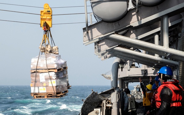 A replenishment-at-sea (RAS) operation is underway between two naval vessels in rough ocean conditions. A pallet of supplies wrapped in plastic is being transferred via a highline/crane system suspended between the ships. Crew members on deck are guiding the load using tag lines while wearing safety gear including hard hats and flotation vests.