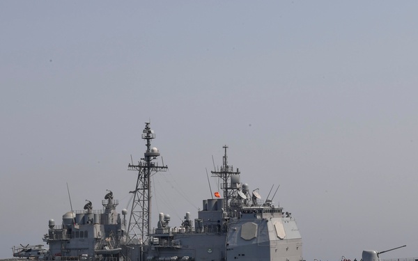 A United States Navy guided-missile cruiser bearing hull number 57 is underway at sea in moderate ocean conditions. The vessel is a Ticonderoga-class cruiser, identifiable by its distinctive superstructure, twin masts, and characteristic gray hull. Personnel are visible on the forward deck near the bow gun mount.
