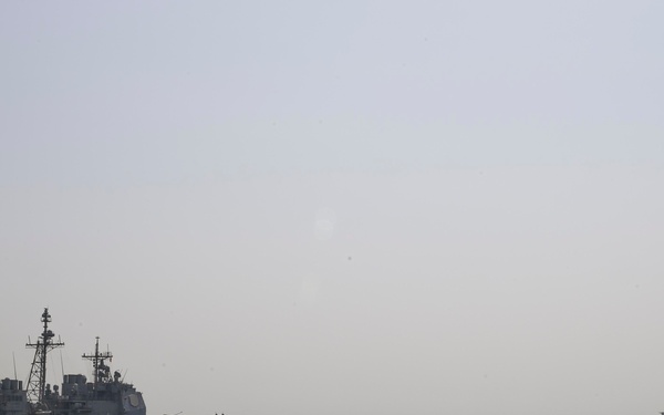The image shows a formation of U.S. Navy warships underway at sea in moderately rough conditions with hazy visibility. At least three vessels are visible, including what appears to be a Ticonderoga-class cruiser or Arleigh Burke-class destroyer in the middle distance, a larger auxiliary/supply ship further in the background, and the bow/superstructure of a warship in the foreground right. Personnel in safety gear are visible on the foreground vessel's deck.