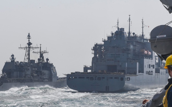 A naval replenishment-at-sea (RAS) operation is underway with multiple vessels in choppy open ocean conditions. Personnel on the foreground vessel, wearing safety vests and hard hats, observe a Ticonderoga-class cruiser and a large auxiliary/replenishment ship operating in close proximity. The sea state appears moderate to rough, with visible wave action between the vessels.