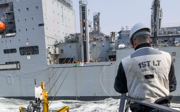 A replenishment-at-sea (RAS) operation is underway between two naval vessels. A pallet of supplies is being transferred via a crane/highline system to the receiving ship's deck. A First Lieutenant from DDG-119 supervises from an elevated platform while crew members in safety gear guide the cargo.