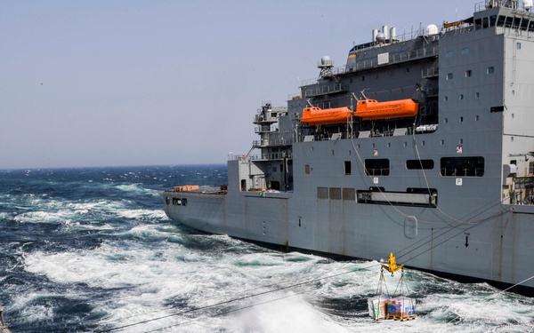 Two naval vessels are conducting an underway replenishment (UNREP) operation at sea, with a cargo pallet suspended on a highline transfer rig between the ships. The larger vessel appears to be a replenishment ship (likely a US Navy auxiliary), while the smaller vessel in the foreground is a warship, possibly a destroyer or cruiser. Rough sea conditions are visible with significant wave action between the two ships.